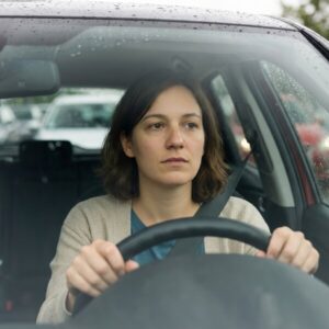 Besorgte Frau am Steuer, Hände am Lenkrad, regennasse Autoscheibe und wartender Verkehr mit Bremslichtern.