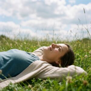Frau liegt entspannt im grünen Gras und blickt lächelnd in den blauen Himmel mit weißen Wolken.