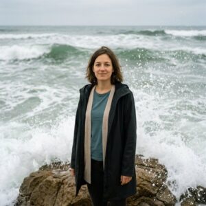 Frau mit geschlossenen Augen, kurzes braunes Haar, beige Strickjacke, steht am Meer, große Wellen schlagen hinter ihr