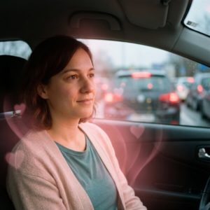 Frau sitzt im Auto und schaut nachdenklich aus dem Fenster; Verkehrsstau im Hintergrund, rosa Herz-Überlagerung