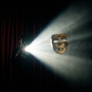 Person am Projektor wirft hellen Lichtstrahl auf schwebende, goldene Theatermaske vor roten Vorhängen