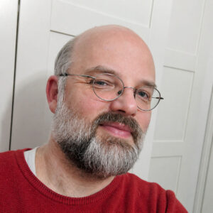 Alexander Rüther mit Brille, Bart und rotem Pullover vor hellem Hintergrund.